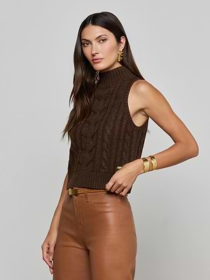 A woman with long brown hair models the L'AGENCE Bellini Cable-Knit Turtleneck, paired with tan leather pants, gold earrings, and a bracelet, posing against a plain light gray background.