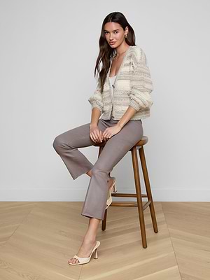 A woman with long brown hair, wearing the Harriet Cardigan with blouson sleeves, taupe cropped pants, and beige heeled sandals, sits on a wooden stool in a minimalist room with light wood floors and white walls.