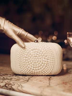 A gloved hand holds the L'Amour Clutch, a handcrafted accessory adorned with small white pearls, resting on marble with a wine glass in the background.