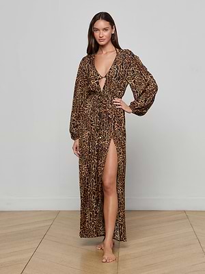 A woman stands barefoot on a wooden floor, wearing the L'AGENCE Sara Maxi Cover-Up Dress featuring a long leopard print design, long sleeves, deep V-neckline, and high slit as she faces forward against a light gray wall.