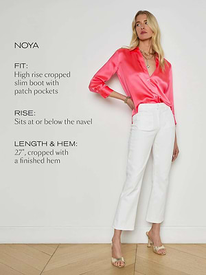 A woman stands against a white wall in the L'AGENCE Noya Cropped Slim Flare Jean, featuring a high-rise and finished hem. She pairs the jeans with a bright pink satin blouse and heeled sandals. Text highlights the pants’ fit, rise, and cropped length.
