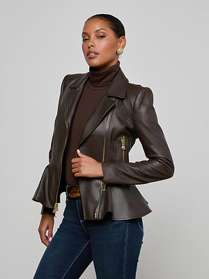A woman with slicked-back hair and gold earrings models the L'AGENCE Lyric Leather Peplum Jacket over a matching turtleneck and blue jeans, standing confidently against a plain light gray background.
