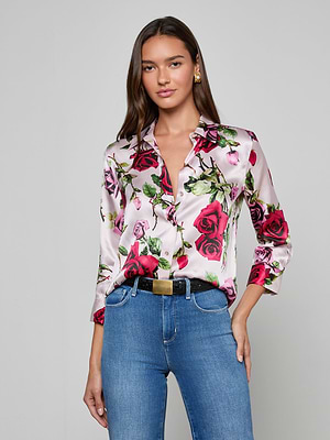 A woman stands against a plain background, wearing the Dani Silk Blouse with a pink and red rose print, blue jeans, a black belt with a gold buckle, and gold earrings.