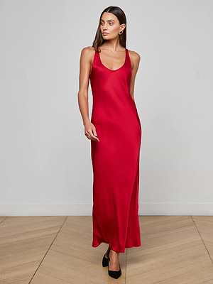 A woman poses indoors against a light wall, wearing the L’AGENCE Akiya Satin Maxi Dress—a sleeveless, floor-length red dress styled with black heels. She has long brown hair and looks slightly to the side.