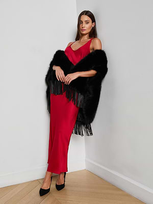 A woman in the L'AGENCE Akiya Satin Maxi Dress and black heels leans against a white wall, draped in a black fringe fur shawl, standing on a wooden floor.