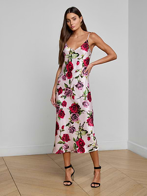 A woman stands indoors on a wooden floor, wearing the Seridie Silk Slip Dress—a white bias-cut slip dress with pink and red rose print—styled with black high-heeled sandals and one hand on her hip.