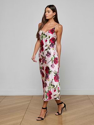 A woman with long brown hair walks on a wooden floor wearing the Seridie Silk Slip Dress in pink with a rose print and black high-heeled sandals. The background is plain and light-colored.