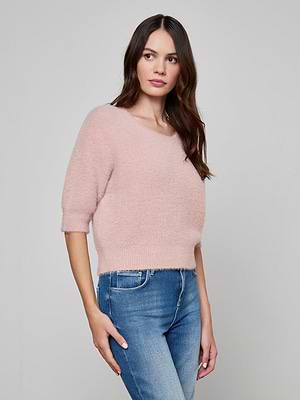 A woman with long brown hair wears the Guinevere Cropped Pullover, an ultra-soft light pink crewneck sweater, paired with blue jeans. She stands against a plain light gray background, looking slightly to her left.